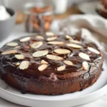 A rich and decadent Queen of Sheba cake with a glossy chocolate glaze, elegantly presented on a white plate with a side of whipped cream and almonds, inspired by Julia Child’s favorite cake.